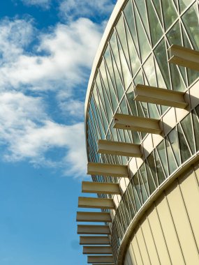 Yapıda metal destekli büyük bir cam dış kaplama yer alıyor. Gökyüzü çoğunlukla dağınık bulutlarla mavidir. Bu görüntü, gündüz saatlerinde bir şehir ortamında binanın bir kısmını gösterir.