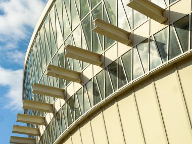 Büyük bir yapı gökyüzünü yansıtan birçok cam pencereye sahiptir. Tasarım, gün içinde bir şehir bölgesinde modern mimarinin altını çizen ön cephedeki farklı unsurları içerir..