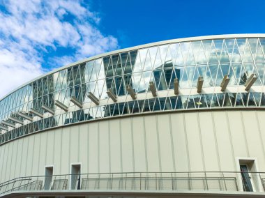 Modern bir binada büyük cam pencereler ve bir açık hava geçidi bulunur. Açık mavi gökyüzü arka planda bulutları gösterir. Yapı şehir ortamında ayakta duruyor..