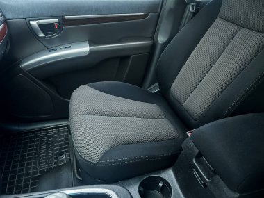 Sahne, bir arabanın sürücü koltuğunu gösteriyor. Koltuğun kumaş bir tasarımı ve modern bir aracın iç dizaynı var. Dışarıdan güzel bir ışık geliyor..