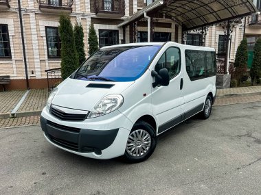 Paraşütü olan bir binanın yanında beyaz bir minibüs var. Minibüs kameraya bakıyor ve şehir merkezinde. Çevrede temiz bir kaldırım ve yeşil ağaçlar var..