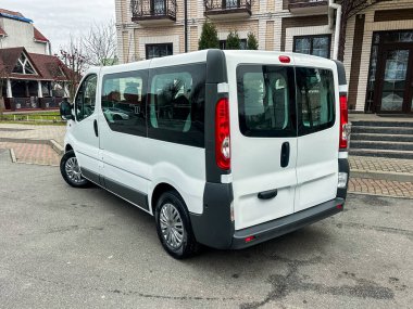 Beyaz bir minibüs tuğla bir yolun yanında bir binanın yanında park halinde. Sahne pürüzsüz kaldırım ve yeşil alanları gösteriyor. Resimde hiç kimse yok..