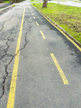 Yol, yolu gösteren sarı çizgileri gösteriyor. Yüzeyde çatlaklar ve yolun yanında çimenler var. Bu bir park alanında gündüz görüntüsü..