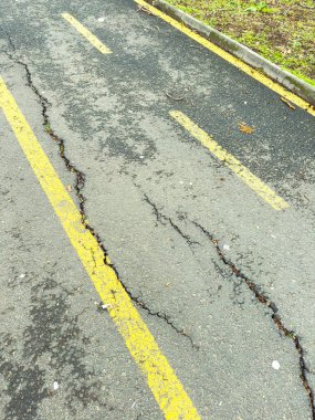 Asfalt yolda çatlaklar görülüyor, sarı çizgiler aşınma izleri gösteriyor. Sahne gün boyunca doğal ışıklandırmayla banliyö bölgesinde geçiyor..