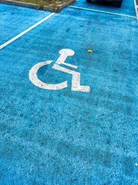 Bir park yeri mavi bir yüzeyde engelli sembolü ile işaretlenir. Arabalar bitişik noktalara park edilmiş. Olay yeri, halka açık bir alanı gün ışığında gösteriyor..