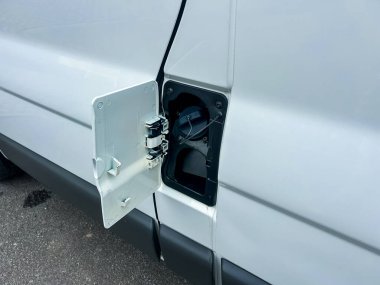 Otoparka park etmiş beyaz bir minibüsün kapağı açık. Gaz çıkışı açıkça görülüyor. Arka planda araçlar var. Sahne bulutlu bir günde çekilmiştir..