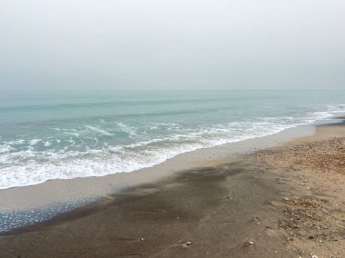 Dalgalar sahile doğru yavaşça akıyor. Kum, su boyunca uzanıyor ve gökyüzü bulutlarla kaplanıyor. Sabahın erken saatleri ve bölge bomboş..