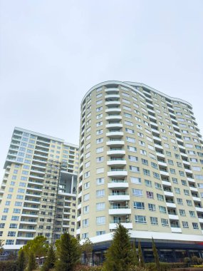 Yoğun bir kentsel alanda bulutlu bir gökyüzüne karşı iki uzun yerleşim yeri yükseliyor. Zemin katta ağaçlar dikilir ve şehirde doğaya bir dokunuş sağlanır..