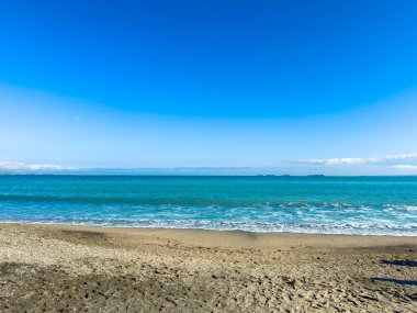 Dalgalar kıyıya yavaşça vururken uzak gemiler açık mavi bir gökyüzünün altında suyun üzerinde yüzüyor. Okyanus kıyısında güneşli bir günde insanlar plajın tadını çıkarırlar..