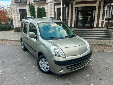 Gümüş renkli bir araba binanın önündeki yolda park halindedir. Ağaçlar yakında ve manzara gökyüzünü gösteriyor. Zaman gündüz vakti..