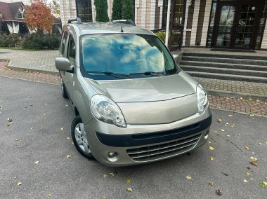 Gümüş renkli bir araba binanın önündeki yolda park halindedir. Gökyüzü açık ve yerde birkaç yaprak var. Sahne, ağaçların olduğu sakin bir bölgeyi gösteriyor..