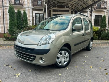 Küçük gri bir minibüs bir binanın önünde park edilmiş, etrafında çatı ve ağaçlar var. Bölgenin temiz bir yolu var ve gündüzleri aydınlık..