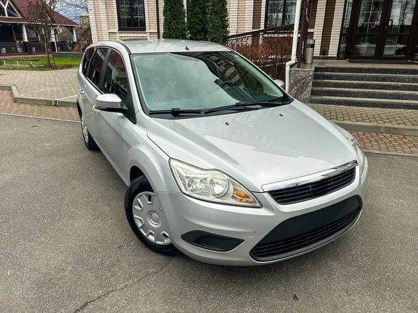 Gümüş renkli bir araba bir binanın önündeki asfalt bir yüzeye park edilmiştir. Gökyüzü açık ve sahne aydınlık. Arka planda yeşil ağaçlar görünür..