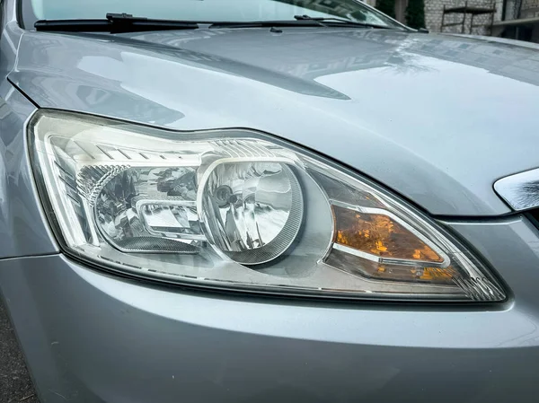 Gümüş renkli bir arabanın farları, araç bir caddede park halindeyken yakına park edilir. Işık net ve görünür, işlevini ve tasarımını gösteriyor..