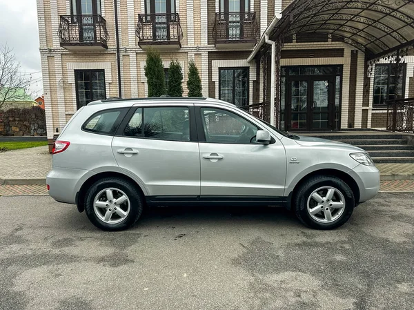 Gümüş renkli bir SUV tuğla duvarlı ve birden fazla balkonlu büyük bir binanın yanına park edilmiş. Zemin asfalt kaplı. Gökyüzü bulutlu, bu da yağmur yağabileceğini gösteriyor..