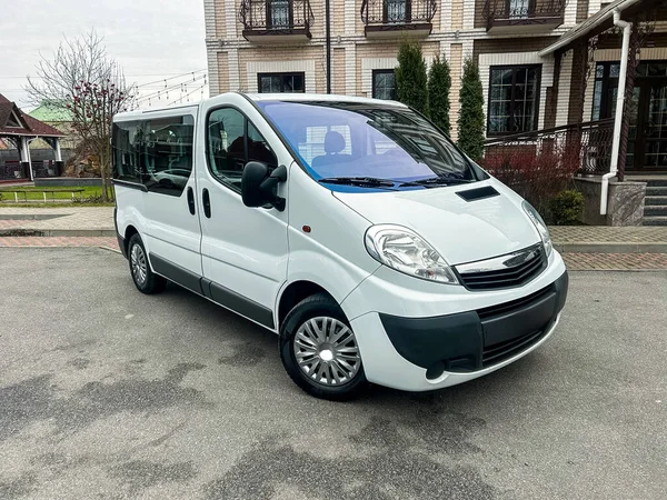 Beyaz bir minibüs büyük bir binanın yanındaki boş bir alana park edilmiş. Bölgede ağaçlar ve yürüyüş yolu var. Güneş ışığı var, aydınlık bir gün..