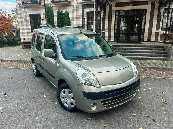 Gümüş renkli bir araba binanın önündeki yolda park halindedir. Ağaçlar yakında ve manzara gökyüzünü gösteriyor. Zaman gündüz vakti..