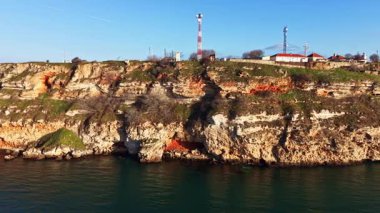 Tepelerinde birkaç iletişim kulesi ve binaları olan uçurumlar su üzerinde yükseliyor. Sahne açık bir gökyüzü ve sakin bir su, sahil boyunca kayalar..
