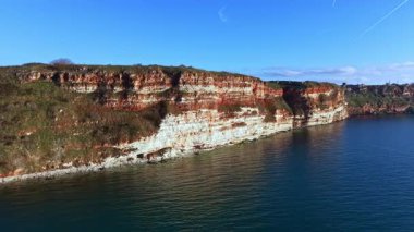 Göze çarpan kayalıkları ve sakin suyu olan kıyı şeridi manzarası. Manzara doğal kaya katmanlarını ve açık mavi gökyüzünü gösteriyor. Bu sahne kıyıdaki doğanın güzelliğini yakalar..