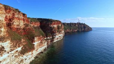 Kızıl ve beyaz kaya katmanlarıyla denizden uçurumlar keskin bir şekilde yükselir. Uzak sahilde bir deniz feneri duruyor. Gökyüzü açık ve mavi. Dalgalar suyun yüzeyinde yavaşça dalgalanır..
