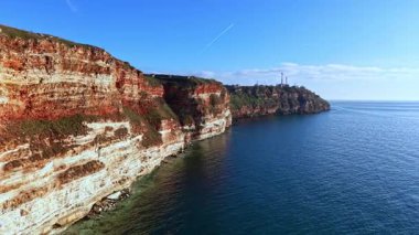 Güneş denizin yanındaki kayalıklarda parlıyor. Dalgalar aşağıdaki kayalara hafifçe dokunur. Açık gökyüzü, kıyı manzarasının güzelliğini artırır..