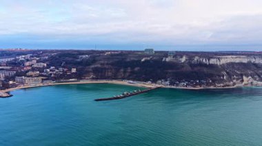 Bir sahil ve liman manzarası, suya demirlemiş tekneleri gösteriyor. Sahilin arkasındaki kayalıklar kıyıda görünen binalarla birlikte yükseliyor. Kışın gökyüzünde bulutlar ve ışık olur..