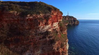 Engebeli kayalıklar mavi suyun üzerinde yükseliyor, doğal manzarayı sergiliyor. Yakın bir yerde açık gökyüzünün altında bir deniz feneri duruyor. Sahne gün ışığında bir sahil ortamı yakalıyor..