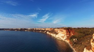 Dalgalar kayalık kıyıya vurduğunda açık gökyüzü mavinin tonlarını gösterir. Kayalıklar suyun üzerinde yükselir ve rüzgar türbinleri uzakta durur. Karadaki suyun yanında küçük bir yapı bulunur..