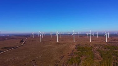 Rüzgar türbinleri açık bir alanda yavaşça döner. Arazi boyunca uzanan toprak bir yol. Parlak mavi gökyüzü hiç bulut göstermiyor. Alanın büyük bölümü ağaçlarla kaplı..