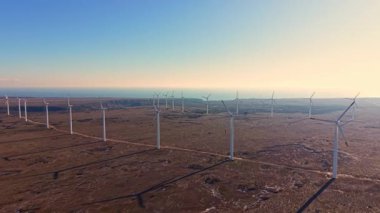 Rüzgar türbinleri gün batımında kıyıya yakın düz arazide uzun bir sıra halinde dururlar. Ufuk açık mavi gökyüzünü ve uzaktaki yumuşak tepeleri gösteriyor..