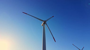Rüzgar türbini, öğleden sonra geç saatlerde mavi gökyüzüne karşı görülebilen kırmızı ve beyaz bıçaklarla yükselir. Sahne yeşil enerji üretimine odaklandığını gösteriyor..