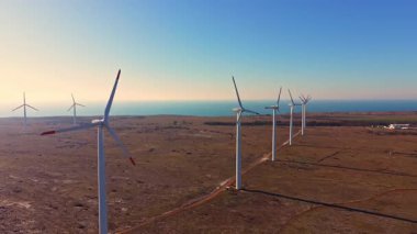 Rüzgâr türbinleri açık bir gökyüzünün altında okyanus yakınında düz bir manzara çiziyor. Rüzgarda dönerek enerji üretirler ve yenilenebilir enerjiye katkıda bulunurlar..