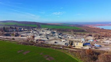 Yoğun bir taş ocağı sahnesi, geniş bir çukurda çalışan büyük makineleri gözler önüne seriyor, yemyeşil tarlalar ve uzak tepelerle çevrili, parlak mavi gökyüzü ve sakin atmosferin altında..