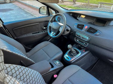 Bir arabanın içinde koltuklar, bir gösterge paneli ve kontroller bulunur. Vites kolu görünür durumda. Güneş ışığı pencerelerden girer, boşluğu aydınlatır..