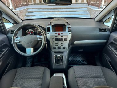 Küçük bir araba binanın önünde park halinde duruyor. Gösterge paneli düğmeler ve kontrollerle görülebilir. Koltuklar kumaştan yapılmış ve kullanım belirtileri gösteriyor. Kar zemini kaplıyor..