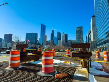 Şikago şehir merkezindeki inşaat bariyerleri, ufuk çizgisi ve mavi gökyüzü bir zemin oluştururken bir bölgeyi kapatıyor. Çalışmalar devam ederken insanlar yakınlarda yürüyebilirler..