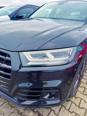 Siyah bir araba çok fazla park edilir. Görüntü, ön taraftaki farları net bir şekilde gösteriyor. Diğer arabalar gün boyunca bulutlu bir gökyüzünün altında park halindedir..