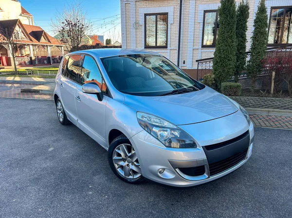 Banliyö bölgesinde beyaz bir evin önüne gümüş renkli bir hatchback araba park edilmiş. Güneş arabanın üzerinde parlıyor ve arka planda ağaçlar görülebiliyor..