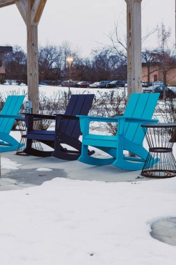 Üç farklı renkte sandalye bir verandaya kurulur ve etrafları karla çevrilidir. Bölge kış belirtileri gösteriyor ve güneş batmaya başlıyor. Çevrede seyrek çalılıklar ve park edilmiş birkaç araba var..