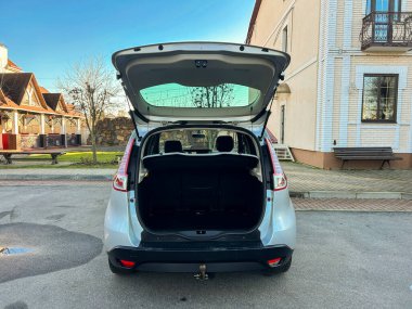 Gümüş renkli bir araba asfalt bir yolda bagajı açık duruyor. Gündüz vakti bir bina ve ağaçlar görünüyor..