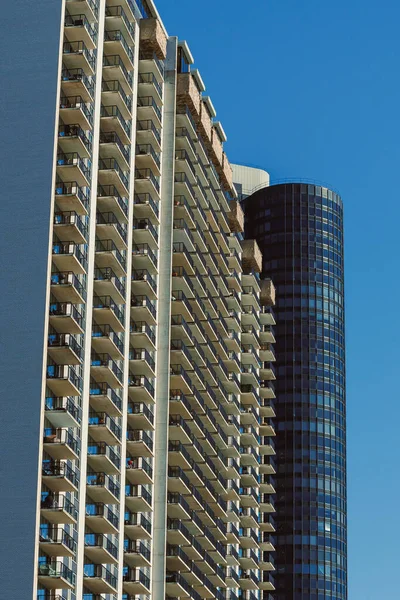 İki yüksek bina açık mavi bir gökyüzünün altında alçak bir açıdan görülür. Balkonları ve cephelerinde kumaşlar var. Güneş ışığı gün boyunca sahneyi aydınlatır..