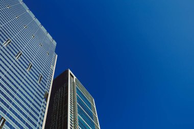 Yoğun bir şehirde iki yüksek bina yan yana durur. Gökyüzü açık mavi ve berrak. Binaların cam pencerelerinde güneş ışığı parlıyor. İnsanlar aşağıda yürür.