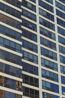 Uzun bir binanın birçok penceresi caddeye bakıyor. Işık camdan yansıyor. İnsanlar aşağıda hareket ederken görülebilir. Yakınlardaki binalar çok hareketli..