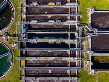 Bir su arıtma tesisinde geniş dairesel tanklar ve uzun boru hatları bulunur. İşçiler yeşil alandaki suyu temizleme ve işletim sistemini yönetmek için mevcuttur.