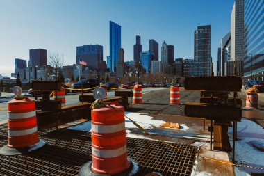İşçiler şehir merkezinde yüksek binaları ve açık mavi gökyüzü olan bir inşaat alanında meşguller. Ön planda bazı yol bariyerleri ve ekipmanlar görülebilir..