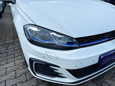 Beyaz bir arabanın önü bir araç parkında park edilmiş. Zemin ıslak ve hava bulutlu. Arabanın parlak farları olan parlak bir tasarımı var..