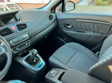 Arabanın içi ön koltukları, gösterge panelini ve vites kollarını gösteriyor. Güneş ışığı pencereden içeri girer. Dışarıda bir park alanı var..