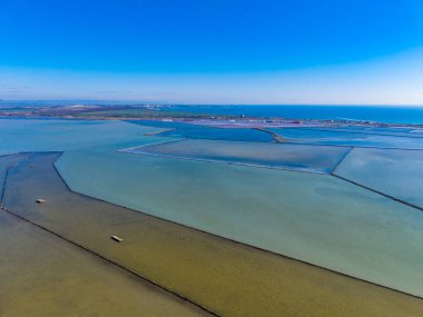 Sahildeki tuz düzlüklerinin havadan görünüşü. Su kanalları manzarada desenler oluşturur. Gökyüzü açık ve mavi. Gün ışığında bölge sular çekilmiştir..