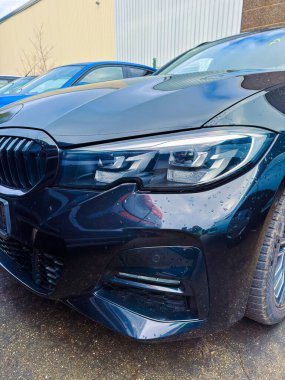 Birçok arabanın arasında siyah bir araba park edilir. Yağmur damlaları yüzeyinde görülebilir. Bulutlu bir gün ve çevrede metal binalar var..