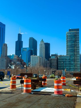 Güneşli bir günde arabalar geçerken, açık mavi gökyüzünün altında arka planda yüksek binalar bulunan bir şehir caddesinde inşaat bariyerleri kuruluyor.